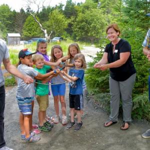 Kids gather around a pair of clippers to help Executive Director Nick Ullo, right, and Environmental Educator Tracey Hall cut the branch and officially open the playground. At left is John Copeland, owner of Kennebec Trail Company which did all the construction of the playground. Peter Southwick photo