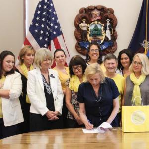 Finding Our Voices survivors and supporters flank Governor Janet T. Mills as she ceremoniously signs a bill initiated by the grassroots nonprofit into law. LD 692 brings safety to women and children survivors of domestic abuse. From left are Adeline and Eva with mom Nicole LeProhon, Jeannine Lauber Oren, Rep. Vicki Doudera, Governor Mills, Eliza Conley Lepene, Sandra Goulet, Dezarae Caron, Dana Ward, Meg Libby, Finding Our Voices founder and president Patrisha McLean, Kerrie McAnulty, Mary Kamradt, and Jennifer Greensmith. Jessi Tracy photo