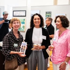 Lucia Droby of Carriage House Gardens, Sherry Dunbar of Tim Dunham Realty, and Francie Siegfried of Birch Home Furnishings & Gifts enjoy the Wiscasset Business After Hours event at the Maine Art Gallery in Wiscasset. In the background, Andy Rogers of Jolie Rogers Raw Bar opens an oyster for Maine Art Gallery Treasurer, John Sawyer. Photo by Bob Bond