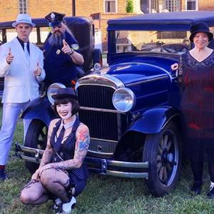 A picture of the cast of Mystery 207's 1920s show. They're dressed like mobsters, flappers, cops, and teetotalers, and pose around a vintage car (maybe a Model T?).